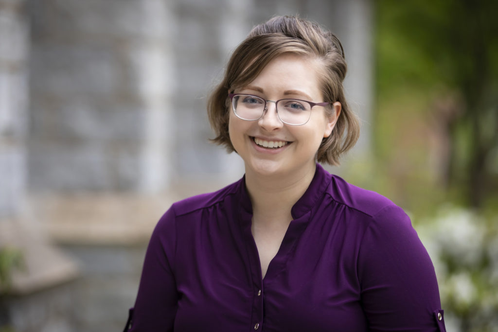 Picture of a white feminine person with short light brown hair, glasses and purple shirt.
